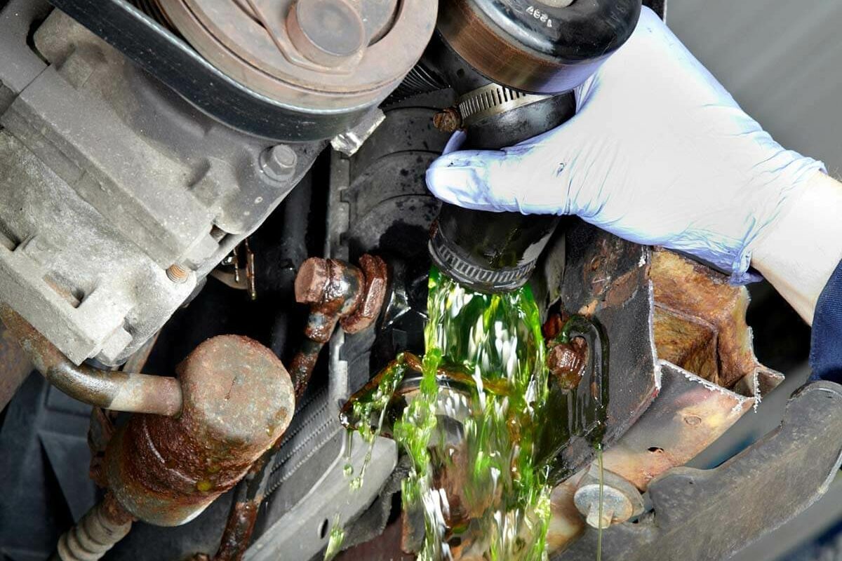 A hand in a glove removes a cap, causing green fluid to spill from a vehicle's engine, surrounded by rusted metal parts and machinery.