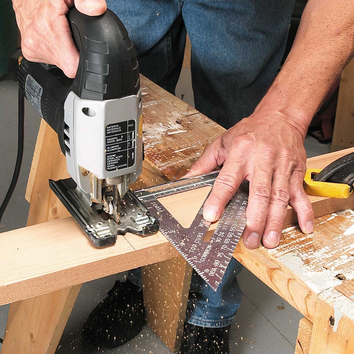A jigsaw cuts through a wooden board while a person holds a metal square to ensure accuracy, surrounded by a workshop with sawdust on the surface.