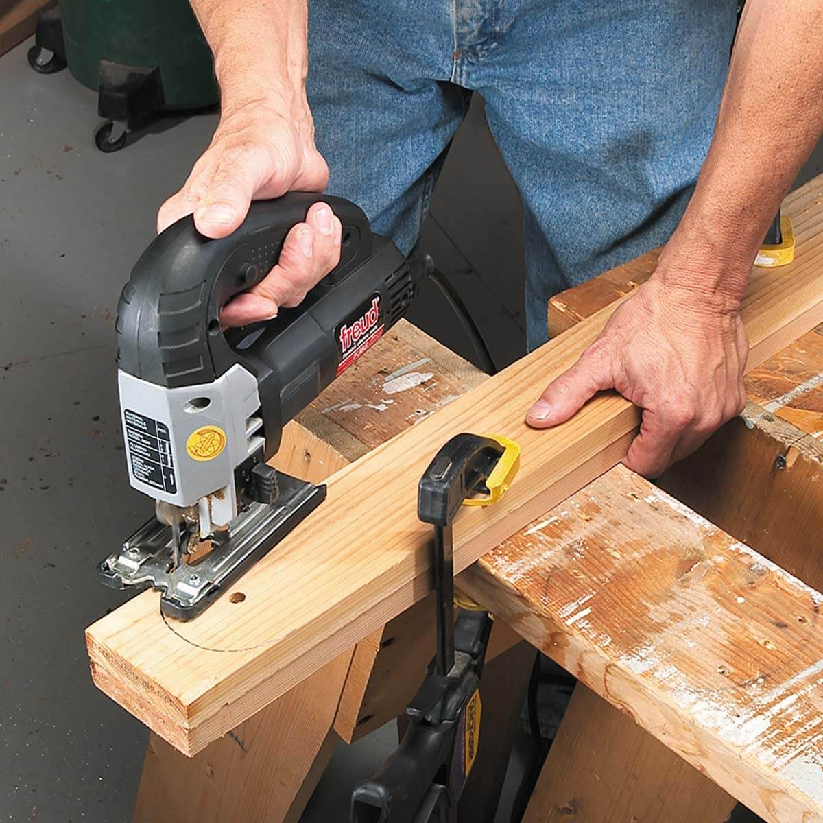 A person uses a jigsaw to cut a wooden plank, securing it to a workbench with clamps in a workshop setting.