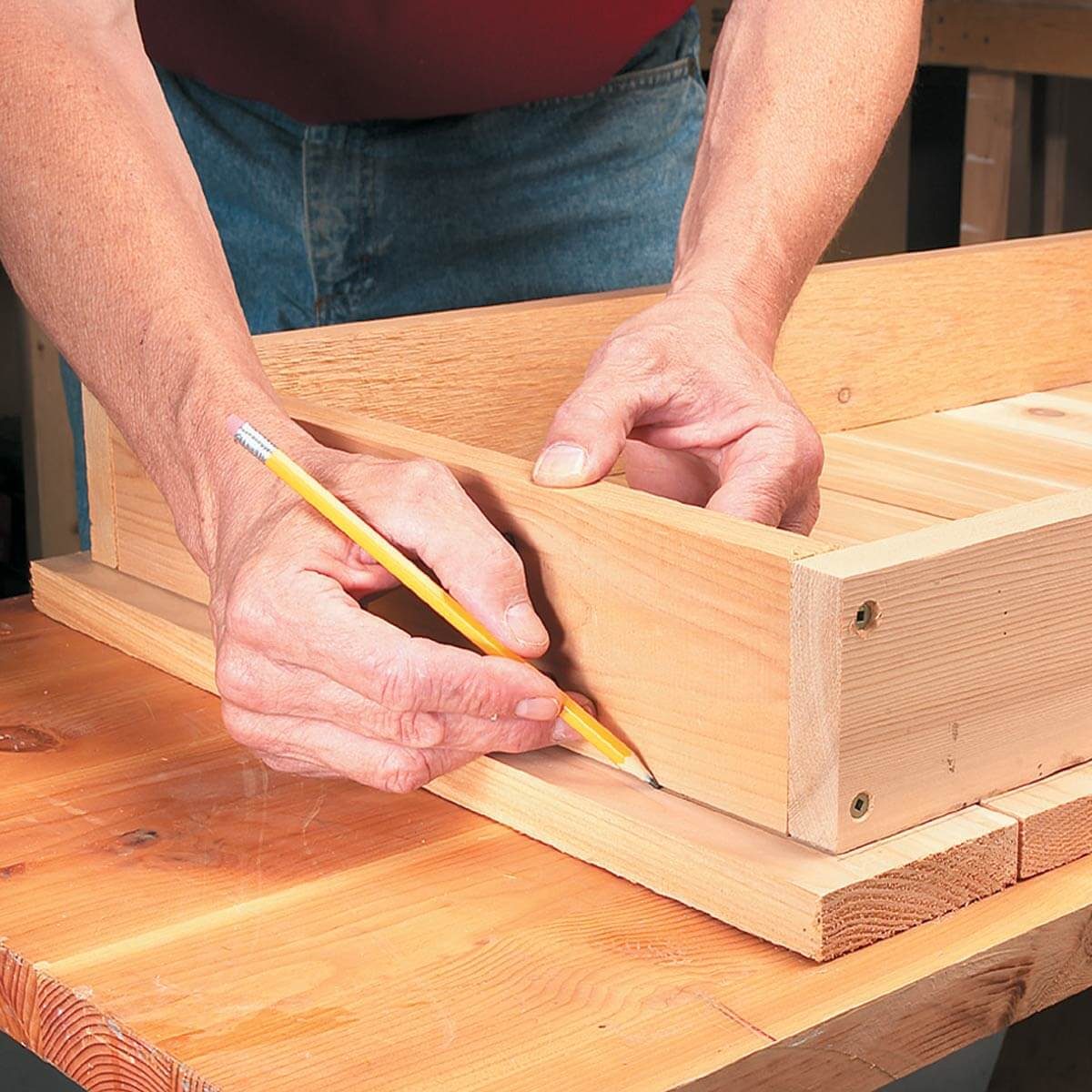 A person holds a pencil, marking a wooden board while positioning another wooden piece. The scene is set on a workbench in a workshop.