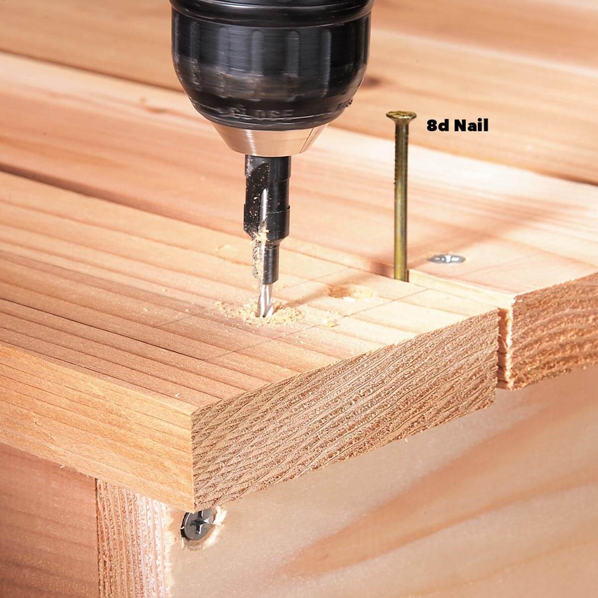 A drill bores into a wooden board, creating shavings. An 8d nail is visible nearby, indicating a construction or woodworking context.