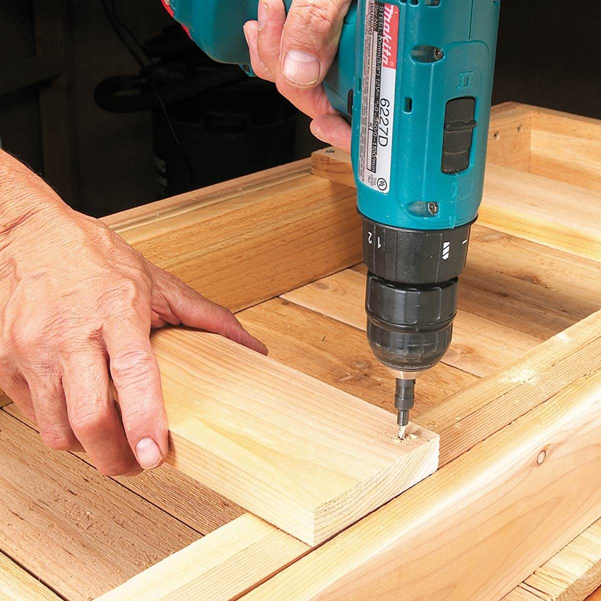 A hand uses a power drill to fasten a wooden plank onto a wooden frame, surrounded by additional wooden pieces and a workshop environment.