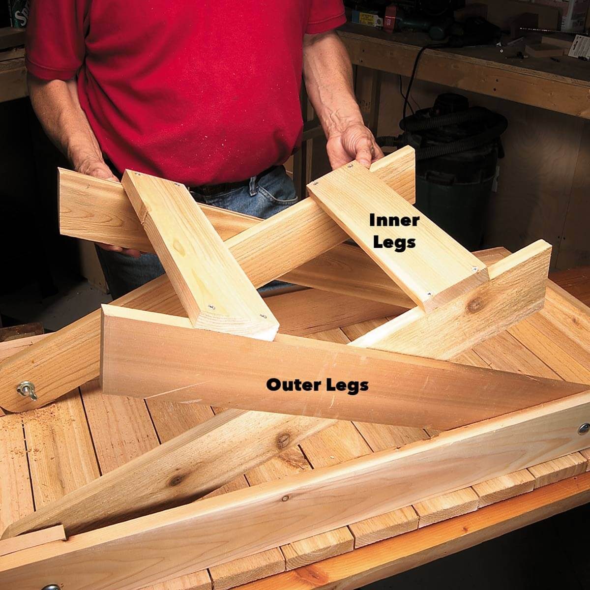 A person holds wooden pieces labeled "Inner Legs" and "Outer Legs," assembling parts on a wooden surface surrounded by tools and materials in a workshop.