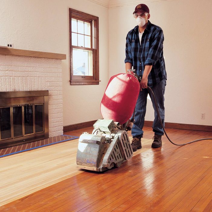 A person uses a floor sander on wooden flooring in a bright room with a fireplace and windows, wearing safety gear and a plaid shirt.