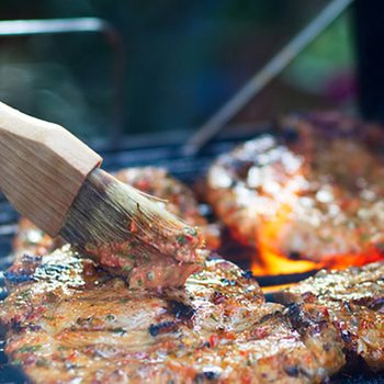 A brush applies marinade to sizzling meat on a grill, surrounded by glowing embers, creating an inviting and aromatic cooking scene.