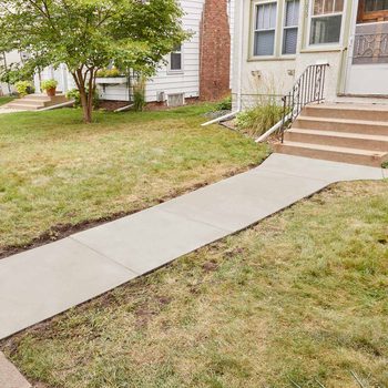 A smooth concrete walkway leads to a set of stairs, surrounded by grass and a small tree, connecting the yard to the front entrance of a house.