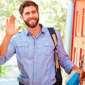 A smiling man waves with one hand while holding a container in the other, standing at a brightly colored door in a green outdoor setting.