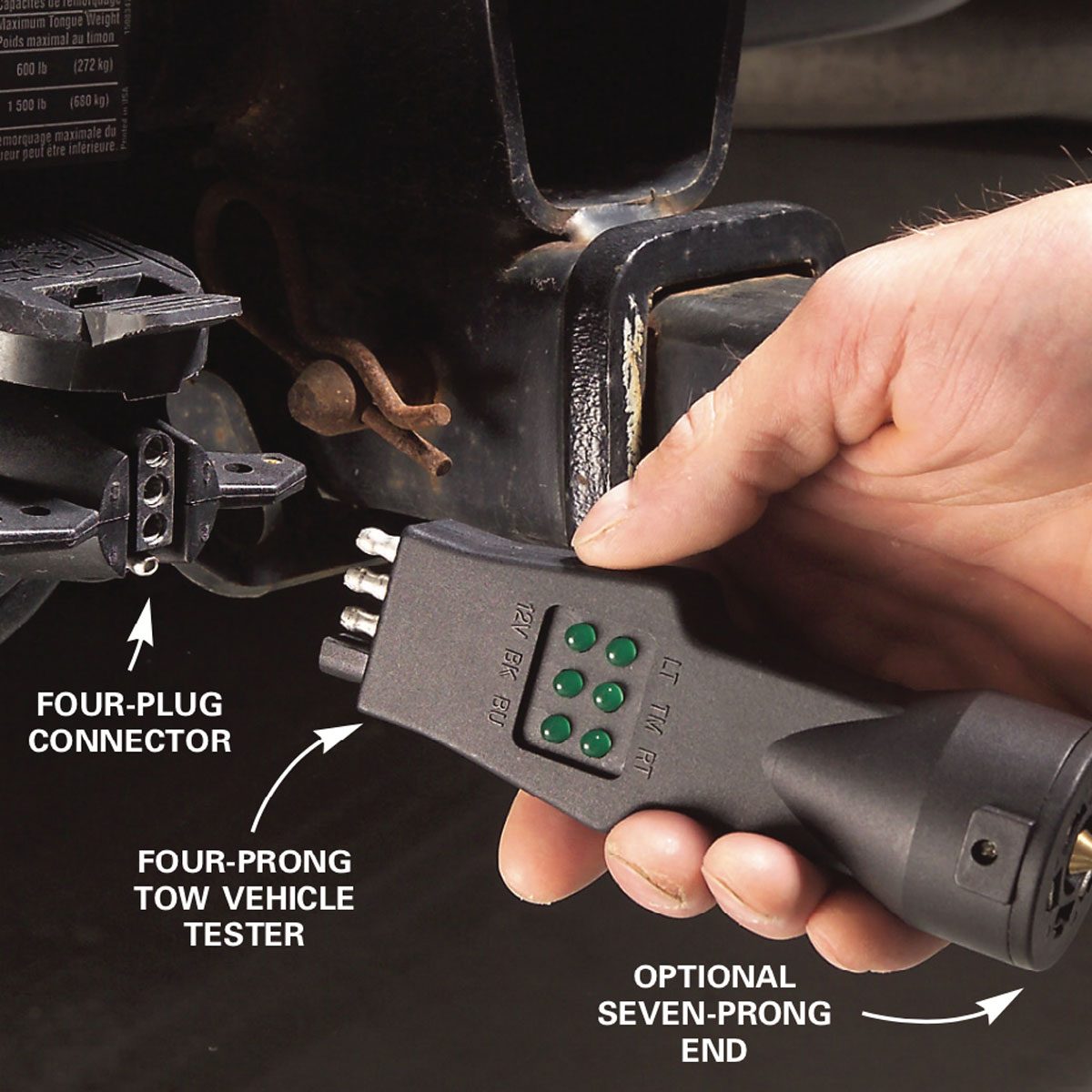 A hand holds a four-prong tow vehicle tester near a four-plug connector, assessing its connection on a hitch in a vehicle's undercarriage.