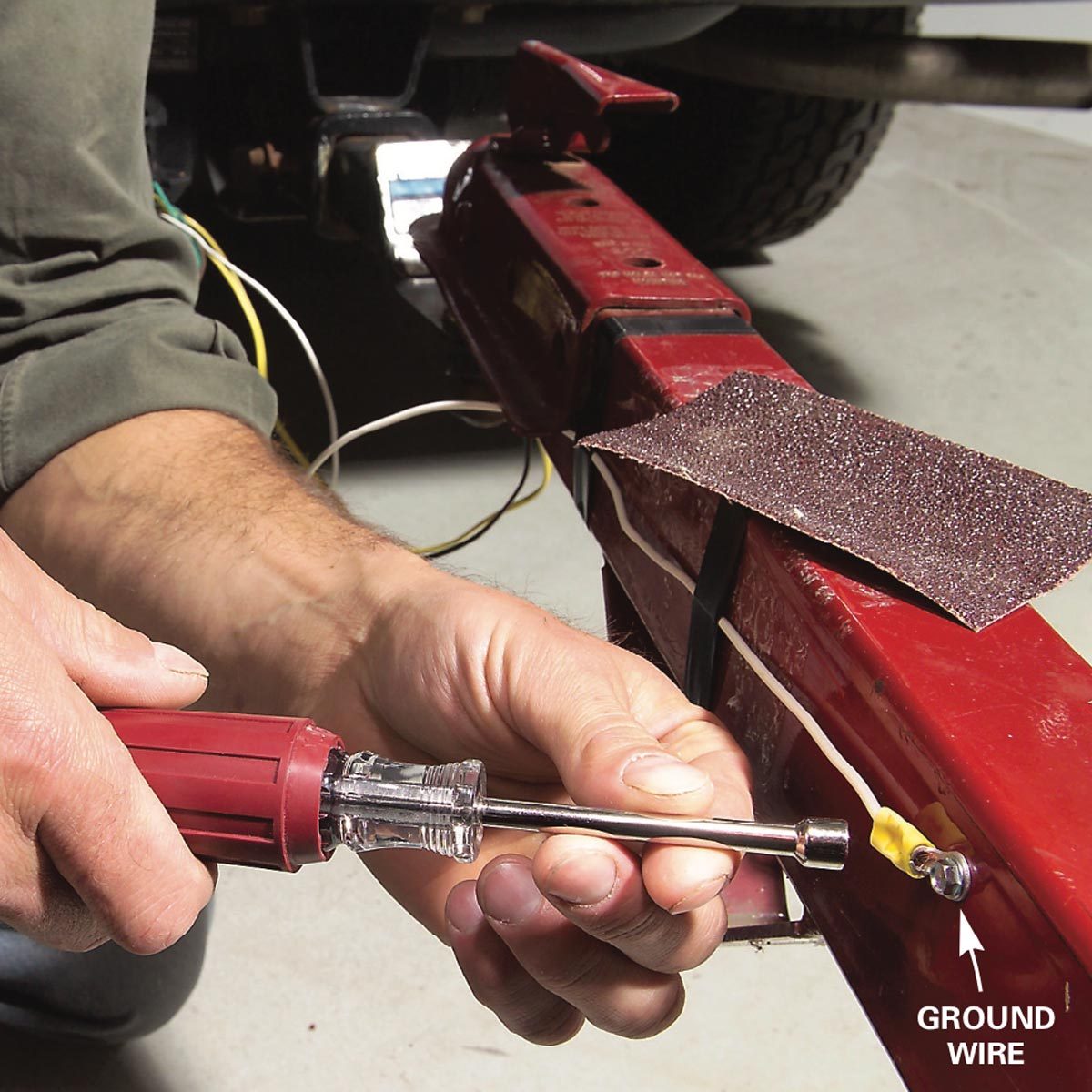 A hand holds a screwdriver, tightening a connection on a red metal frame, with yellow wire and sandpaper nearby in a garage setting.