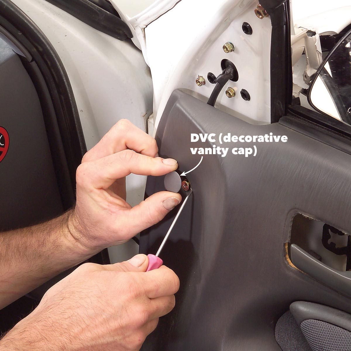 A hand is using a tool to remove a decorative vanity cap (DVC) from a vehicle's interior panel, surrounded by a car door's components.