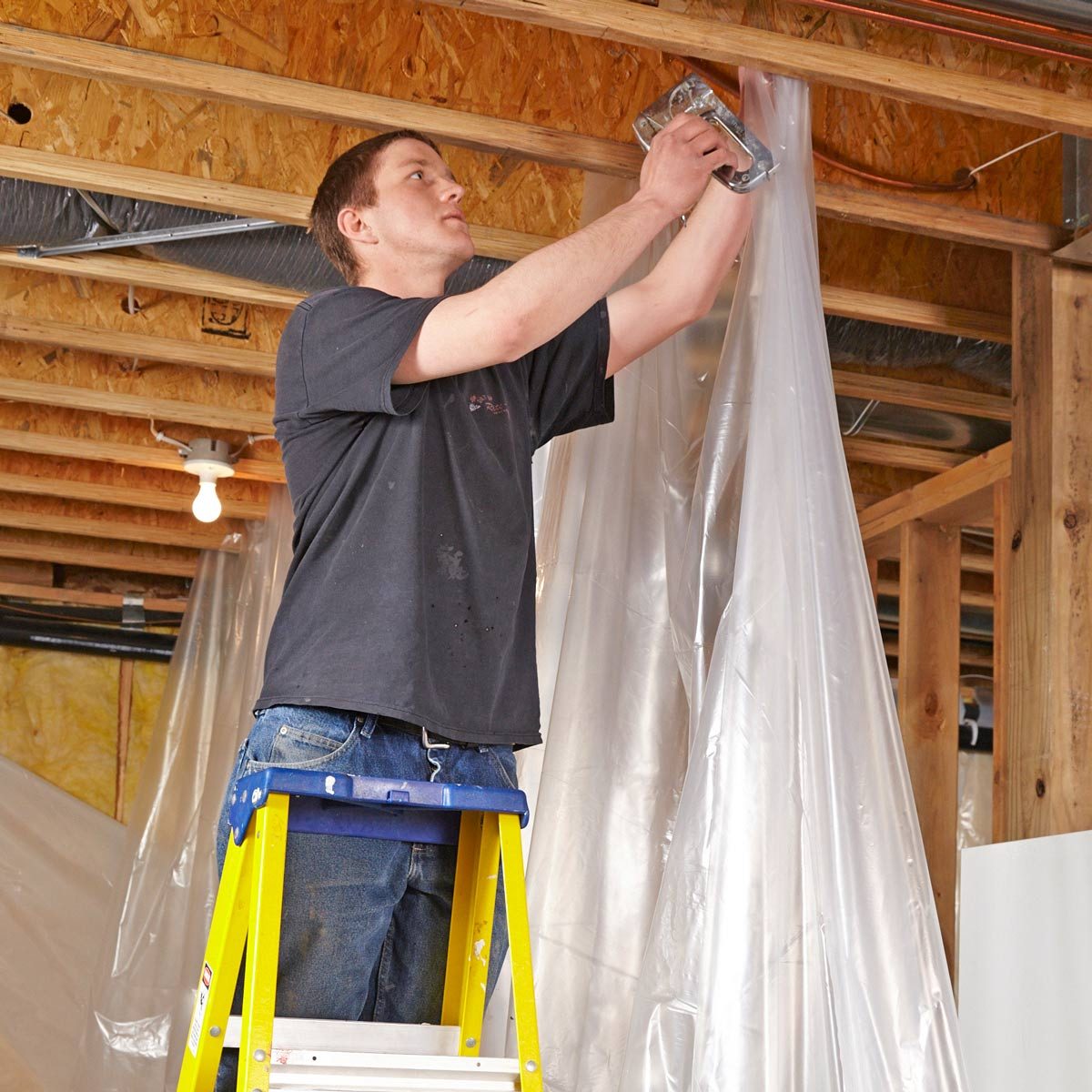 A man stands on a yellow ladder, attaching plastic sheeting to a wooden ceiling framework in a construction setting with exposed beams and a bare bulb.