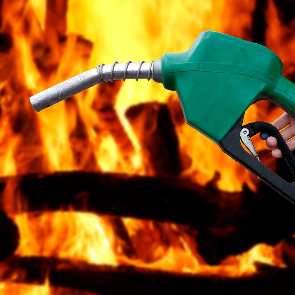 A green fuel nozzle is held over roaring flames, suggesting an action that may involve refueling or igniting a fire. Logs are visible in the background.