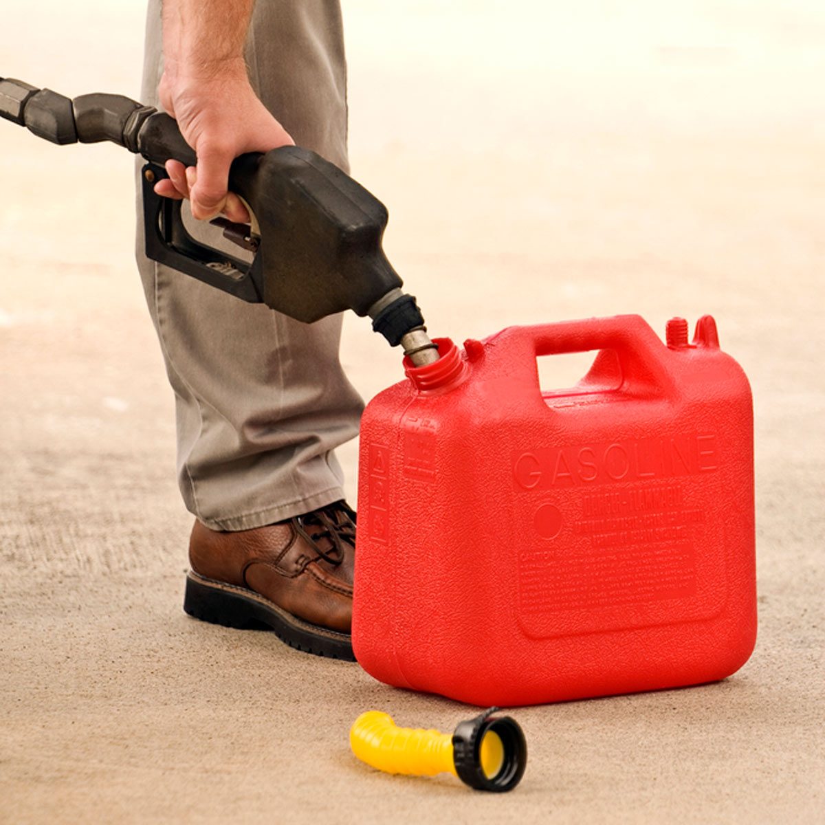 A person pours gasoline from a nozzle into a red gasoline canister, situated on a concrete surface, while wearing brown shoes.