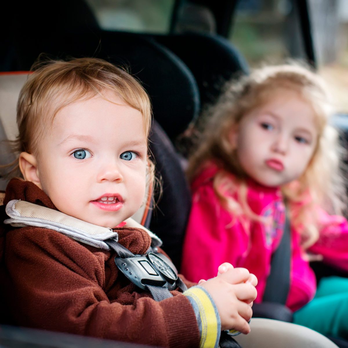 A toddler in a car seat smiles, while an older child pouts in the background, both inside a vehicle. They are seated in a cozy, safe environment.