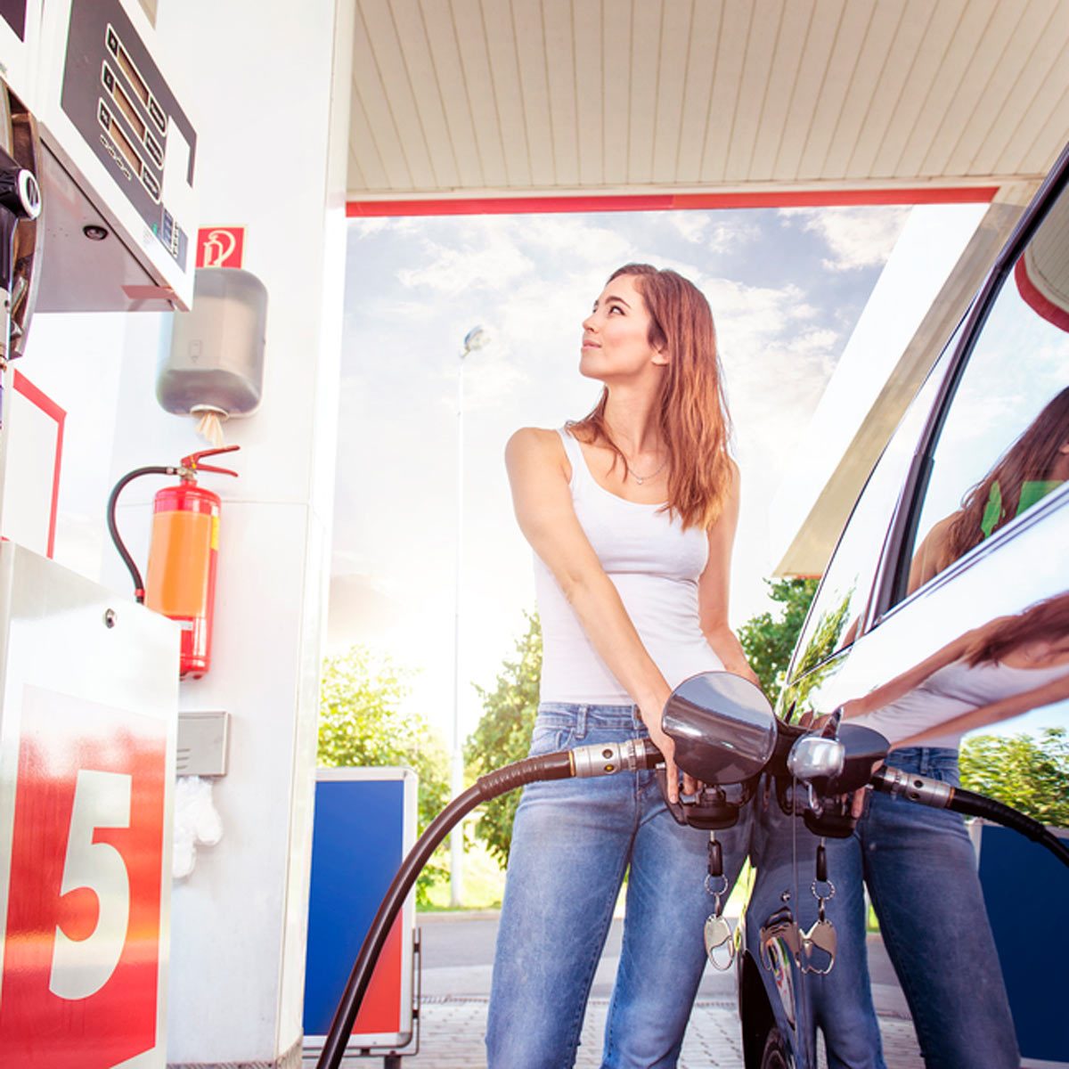 A woman is fueling a car at a gas station, looking up and smiling, surrounded by vehicles and greenery under a clear sky.