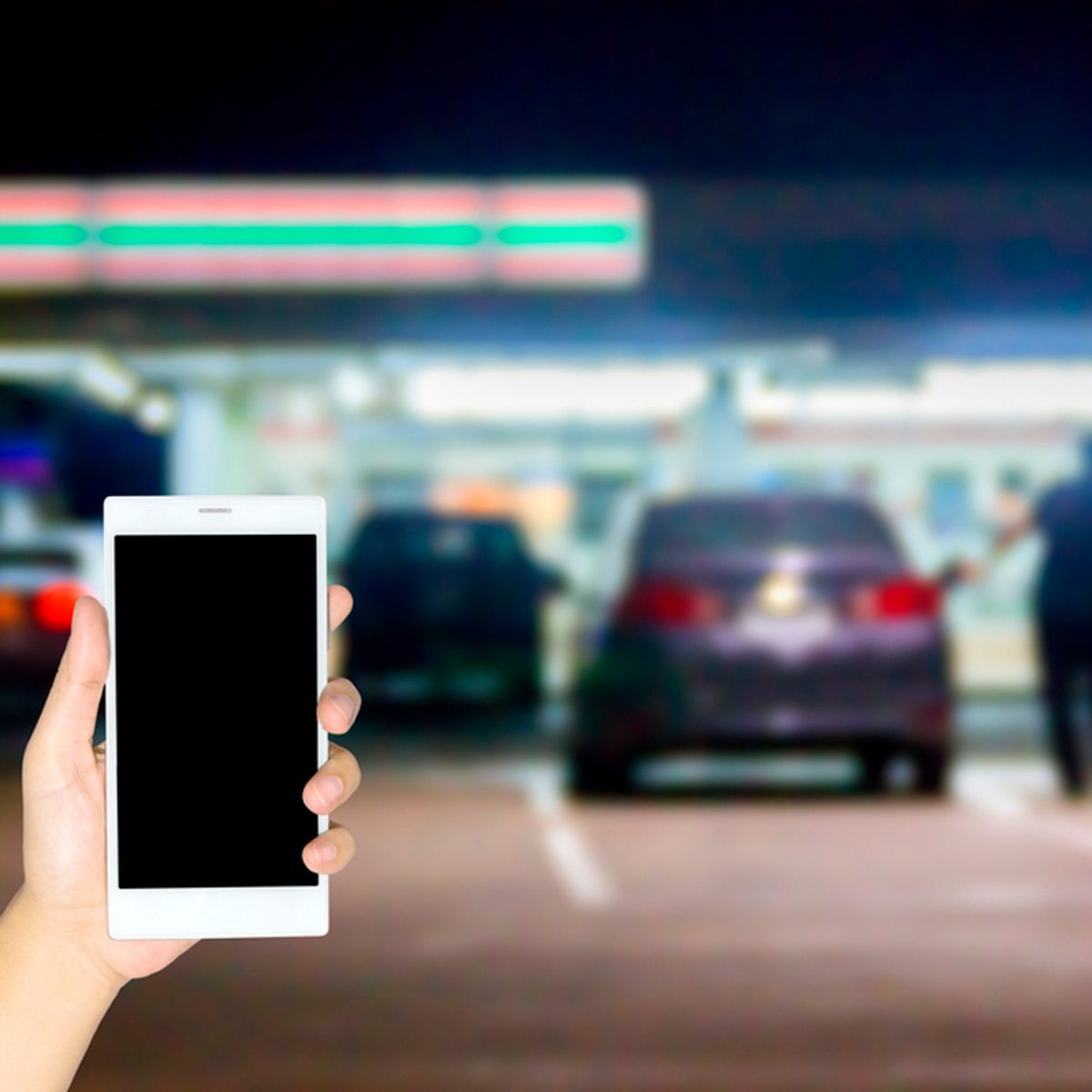 A hand holds a smartphone, while blurred cars are parked in a dimly lit area, suggesting a nighttime setting near a gas station or convenience store.