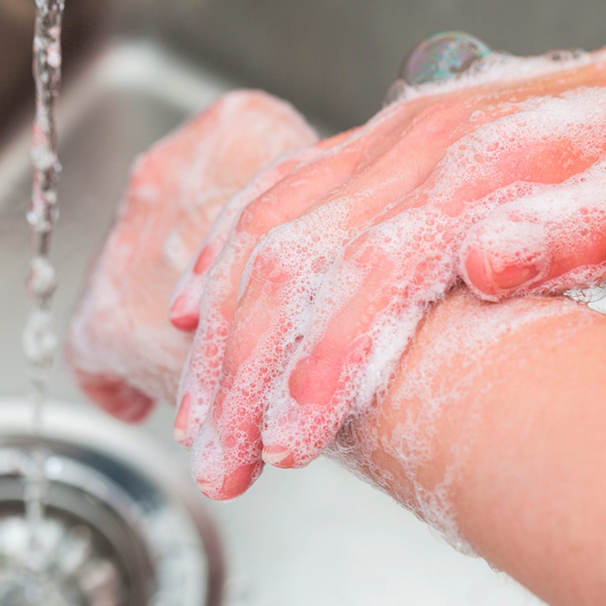 Soapy hands are being scrubbed under running water at a sink, with a shiny drain visible below.