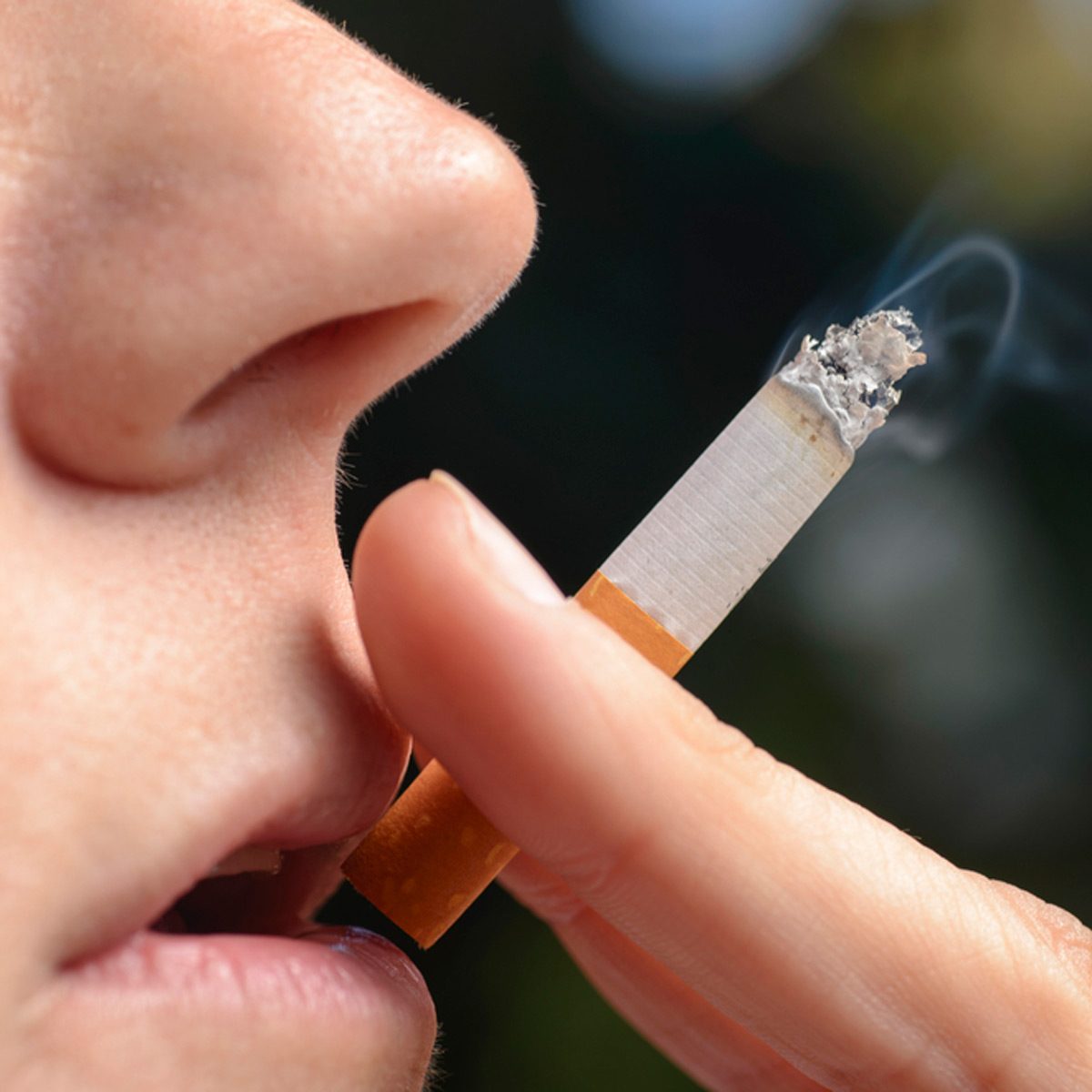 A hand holds a cigarette near a person's lips, as smoke rises from the burning end, set against a blurred outdoor background.