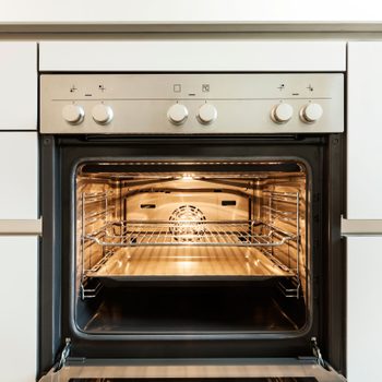An oven is open, with a visible light inside, showcasing metal racks and a baking tray in a modern kitchen setting.