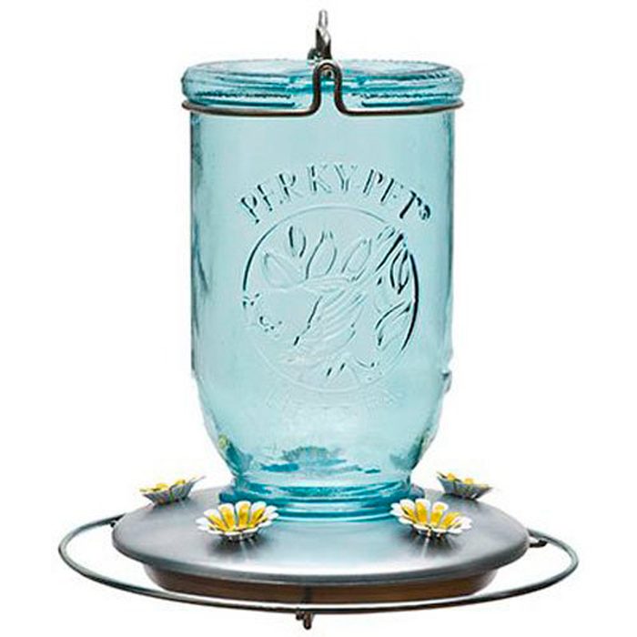 A clear blue glass hummingbird feeder hangs, with a metal base featuring flower-shaped feeding ports, designed to attract hummingbirds in an outdoor setting.