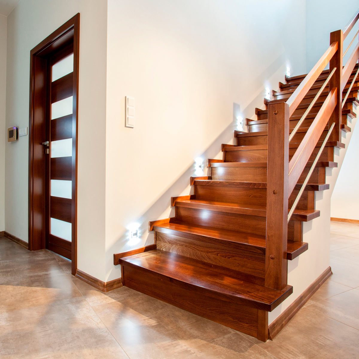Wooden stairs ascend with bright lights beside them, leading to an open space. A modern door and light-colored walls frame the area.