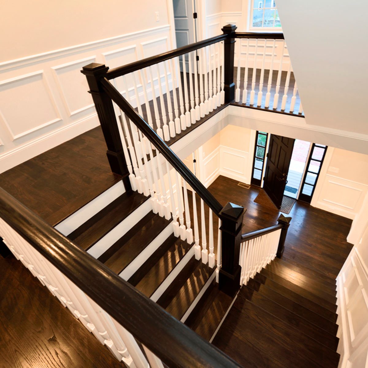 Wooden stairs curve downwards with white railings, leading into a well-lit foyer featuring a dark door and large windows, amidst light-colored walls.