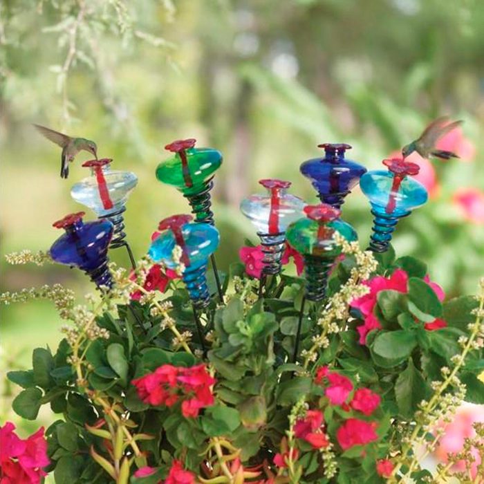 Colorful glass flowers attract a hummingbird, surrounded by green foliage and blooming pink flowers in a vibrant outdoor garden setting.