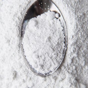 A silver spoon holds white powder, surrounded by a larger pile of powder on a smooth, light-colored surface, creating a clean and minimalistic appearance.