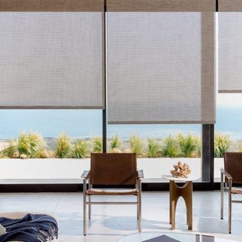 Three modern chairs sit on a tiled floor, facing large windows with blinds, revealing a view of greenery and ocean beyond.