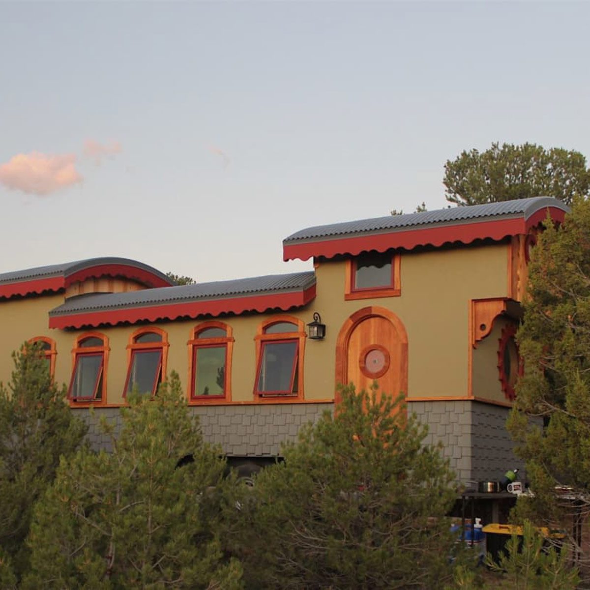 A colorful, uniquely shaped house sits elevated on stilts among green trees, featuring large windows and decorative wooden accents under a clear sky.