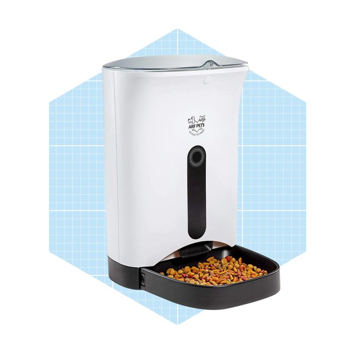A pet food dispenser fills an attached bowl with kibble, placed against a light blue grid background. It features a sleek white design with a transparent lid.
