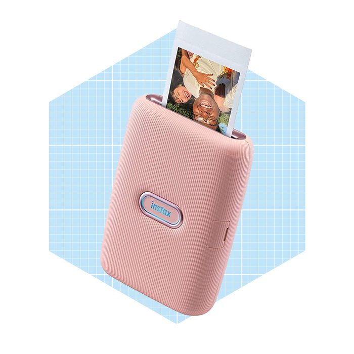 A pink instant photo printer is ejecting a printed image in a grid-patterned blue background. The visible photo shows two smiling people.