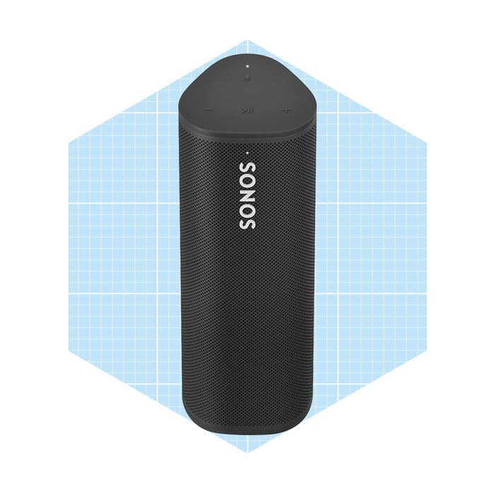 A black portable speaker stands upright, showcasing control buttons on top, against a light blue grid-patterned background. The brand name 
