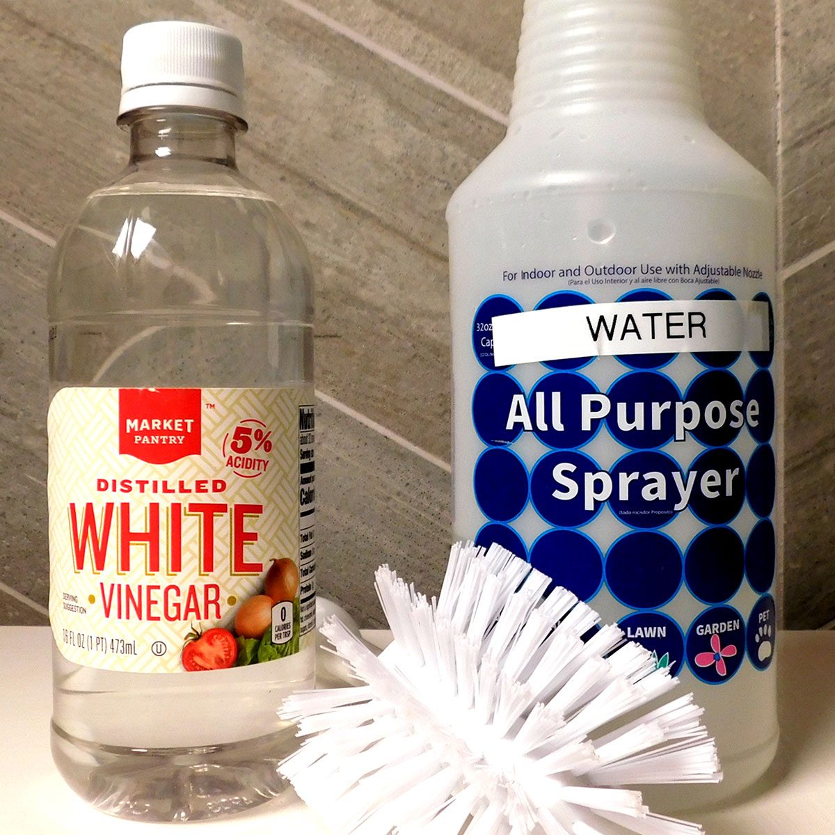 A bottle of white vinegar and an all-purpose spray bottle sit next to a cleaning brush on a tiled surface, likely in a kitchen or bathroom.