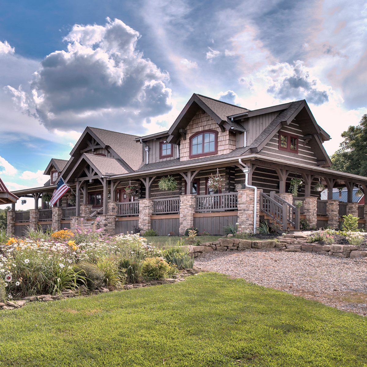 A large, two-story house with a wooden and stone facade stands amidst lush green grass and colorful flower beds under a cloudy sky.