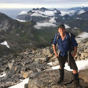 A man stands on rocky terrain, smiling and wearing hiking gear, with towering mountains and clouds in the background under a gray sky.