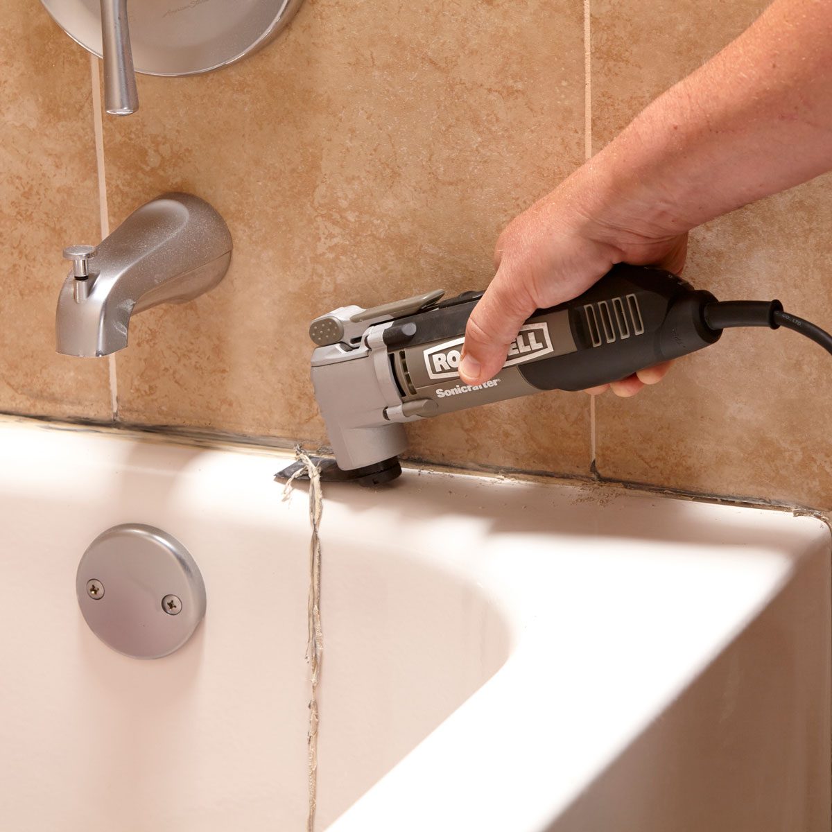 A tool held by a hand is removing old caulk from a bathtub's edge near a faucet, surrounded by tiled walls and a circular drain cover.