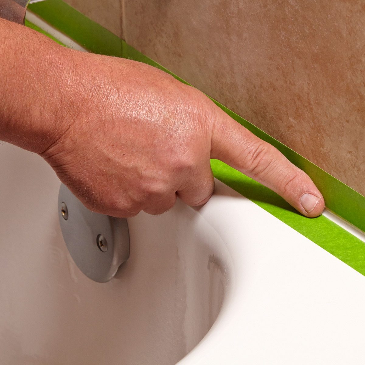 A hand presses down masking tape along the edge of a bathtub, aligning it with a tiled wall in a bathroom renovation setting.