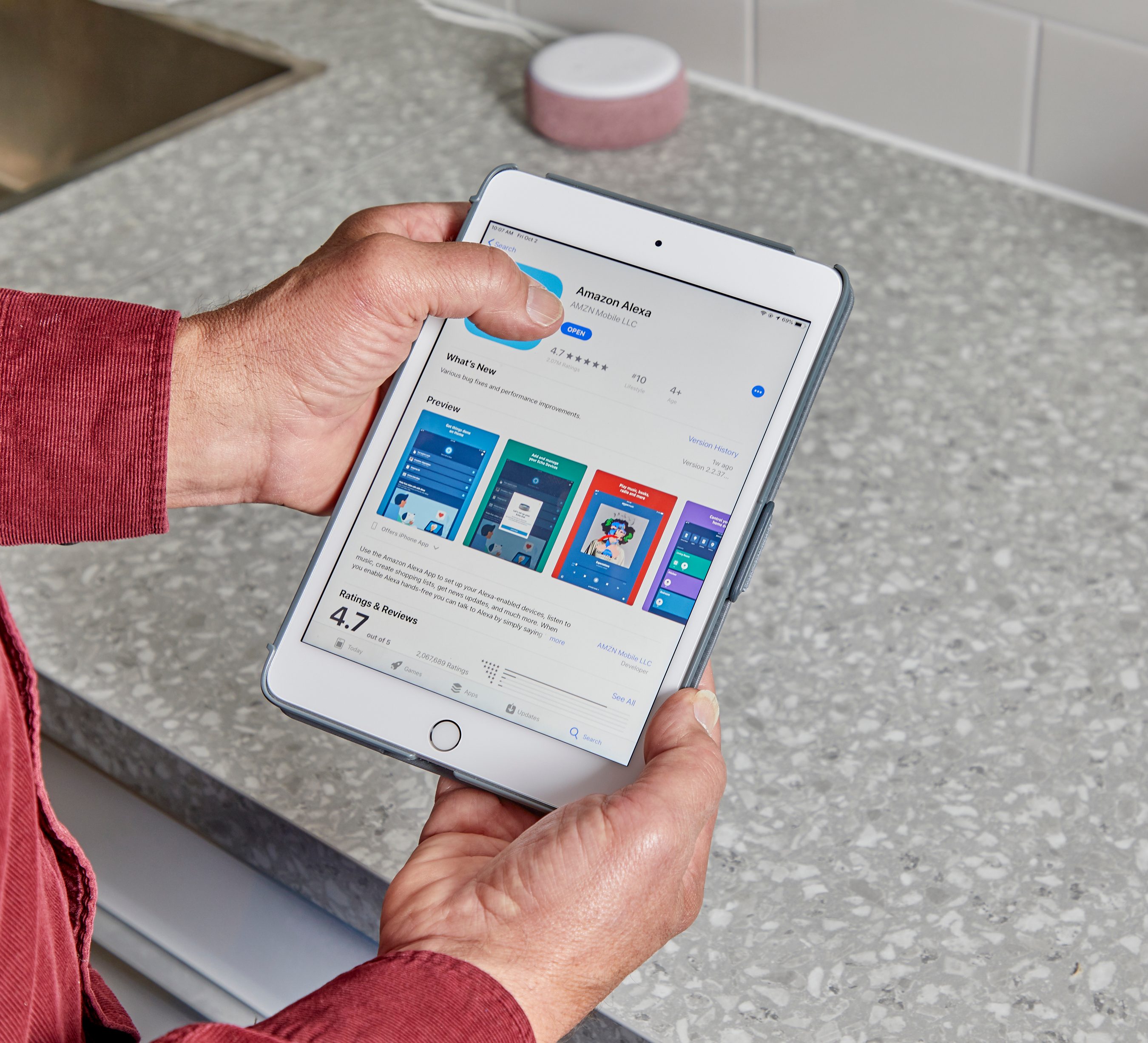 A person holds an iPad displaying the Amazon Alexa app, while standing near a counter with a sink and a round object.