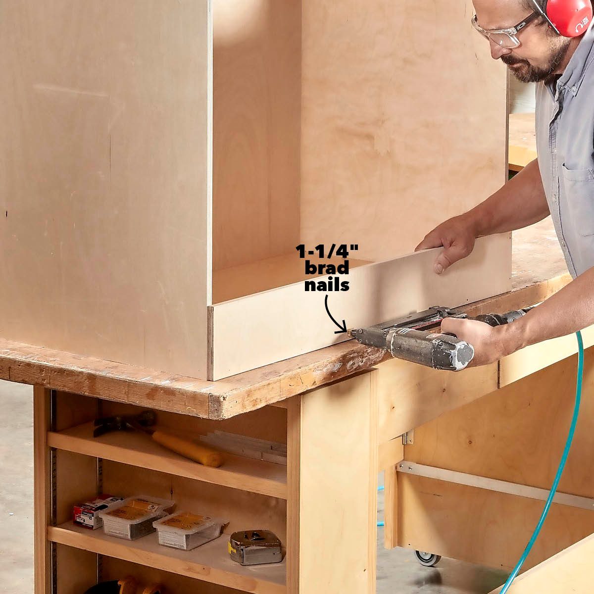 A person uses a brad nailer to attach a panel to a woodworking project on a table, with tools neatly arranged below.