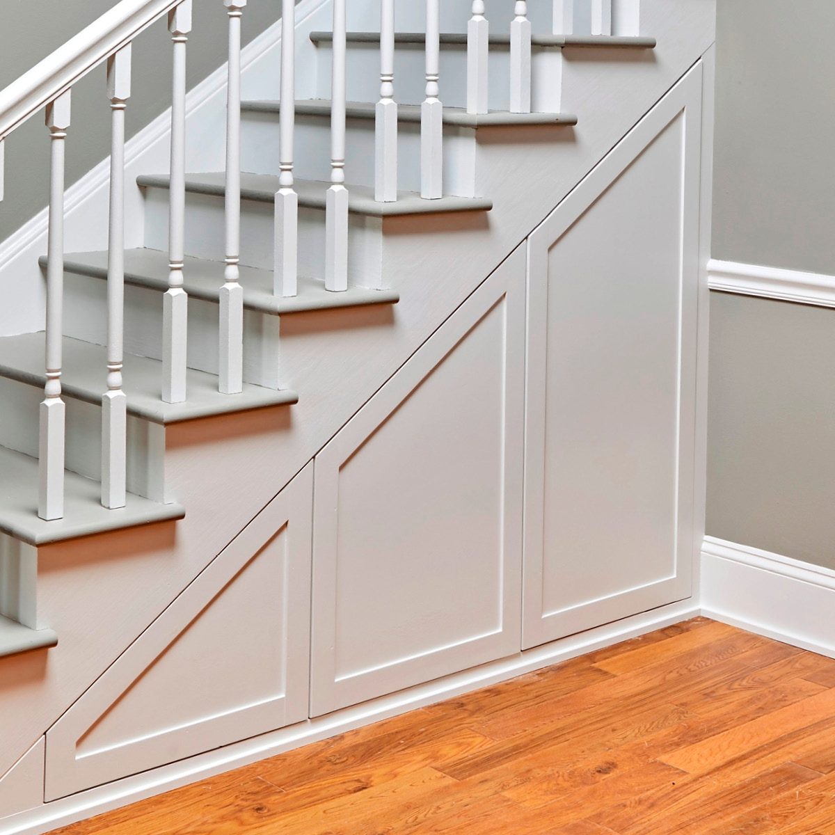A white staircase ascends, with wooden steps and railings. The area beneath the stairs features paneled storage, against a light gray wall and hardwood floor.