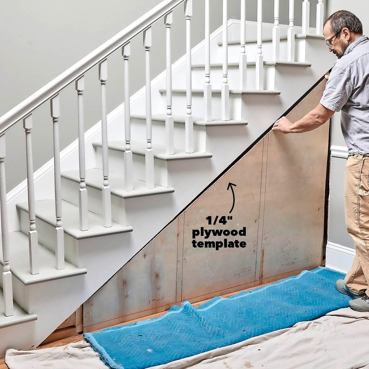 A man measures a plywood template against the wall under a staircase, surrounded by a light-colored interior and blue protective covering on the floor.