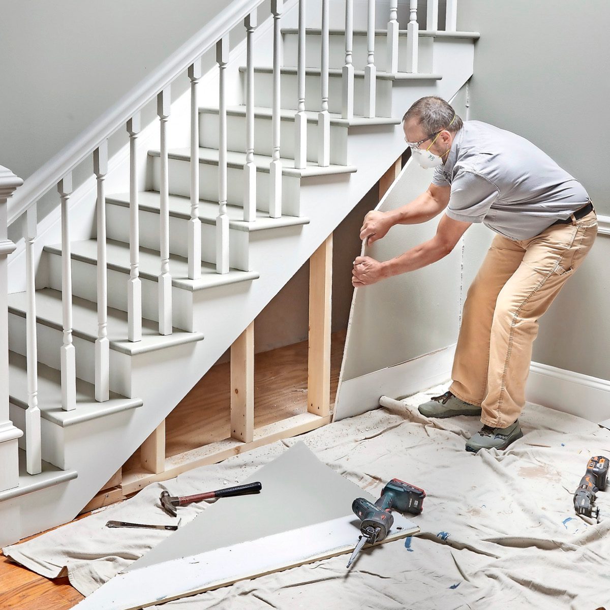A man is installing drywall under a staircase while using tools on a tarp-covered floor in a well-lit indoor space.