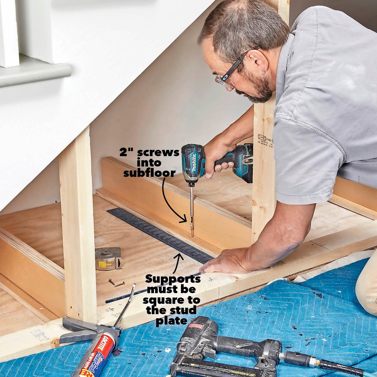 A contractor uses a power drill to secure a wooden support to a subfloor, surrounded by tools and materials on a protective blue mat.