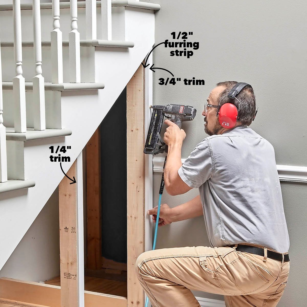 A man uses a nail gun to attach trim and furring strips under a stairwell, in a room with green walls and white molding.