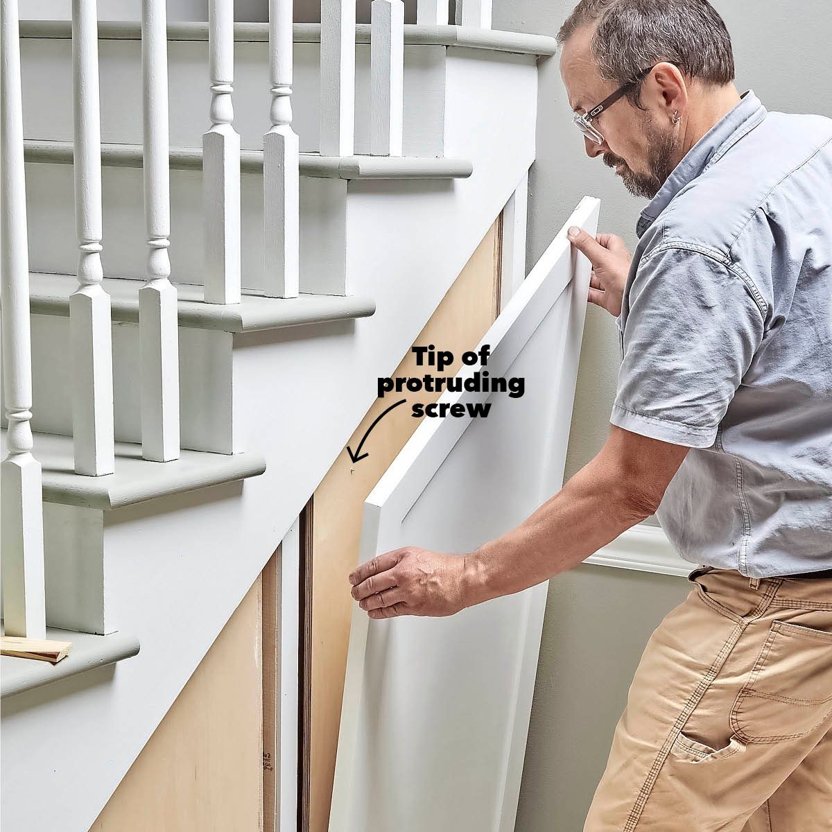 A person is positioning a panel near the base of a staircase, while a screw tip is visibly protruding from the wall beside them.