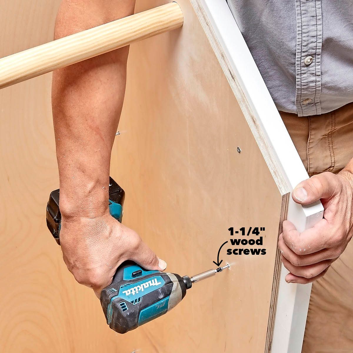 A person is using a power drill to attach a white panel to a wooden structure, with focus on inserting a 1-1/4" wood screw.