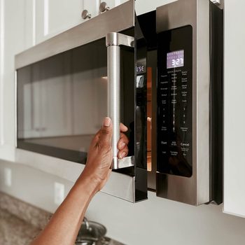 A hand pulls open a stainless steel microwave door, with a digital display showing the time and several control buttons, set against a kitchen background.
