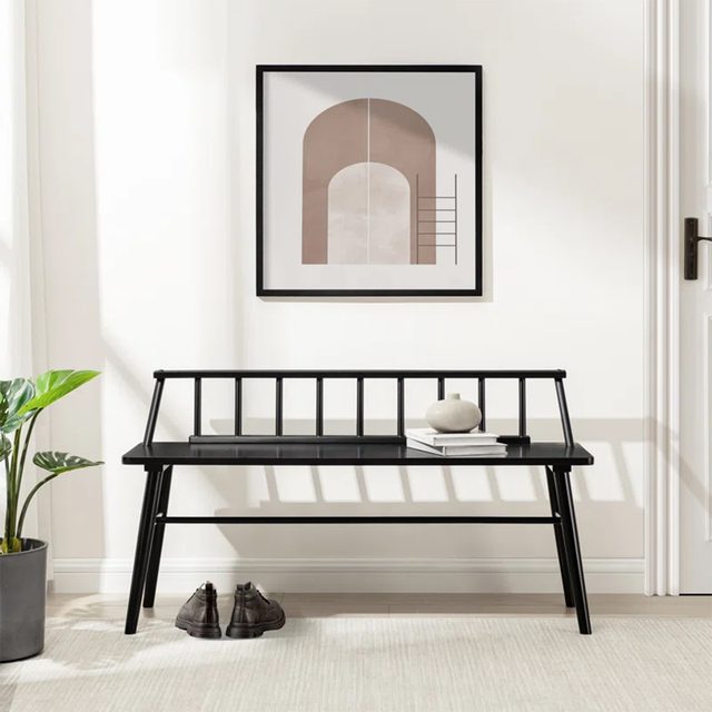 A black wooden bench sits against a bright wall, with a geometric artwork above it and a potted plant nearby, creating a minimalist interior setting.