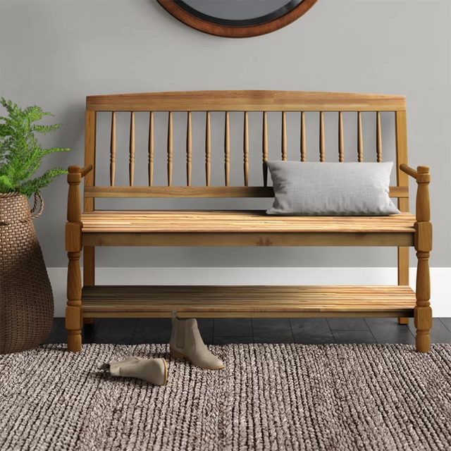 A wooden bench sits against a gray wall, displaying a gray pillow, while casual boots lie on a textured rug nearby. A potted plant adds greenery.